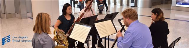 Picture of small orchestra performing in one of the terminals, the instruments include; Clarinet, bassoon, flute, oboe, and french horn. 